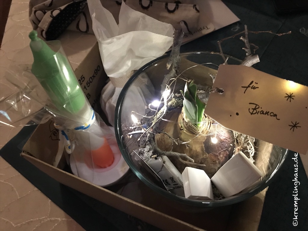 Geschenkbox mit grün-weiß-orange farbener Kerze in weißem Kerzenständer, Amarylis in Glas mit Lichterkette und tüte mit Plätzchen